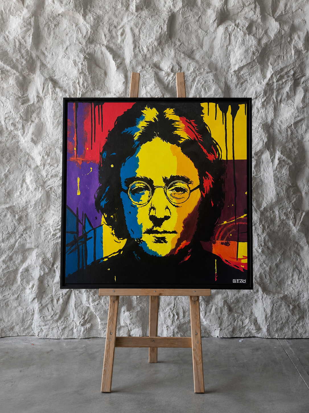 A framed pop-art portrait print of John Lennon displayed on a wooden easel against a textured light gray wall and concrete floor.